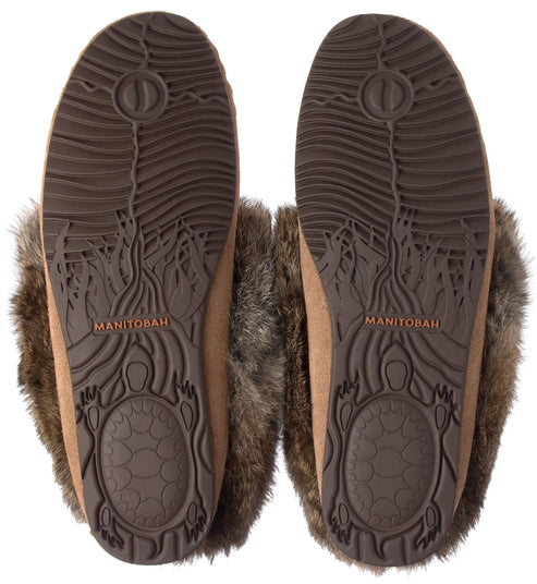 A pair of tan suede slippers with dark brown faux fur trim, shown from the bottom to display the molded rubber soles featuring a turtle graphic, a wave-like pattern, and the embossed name MANITOBAH.