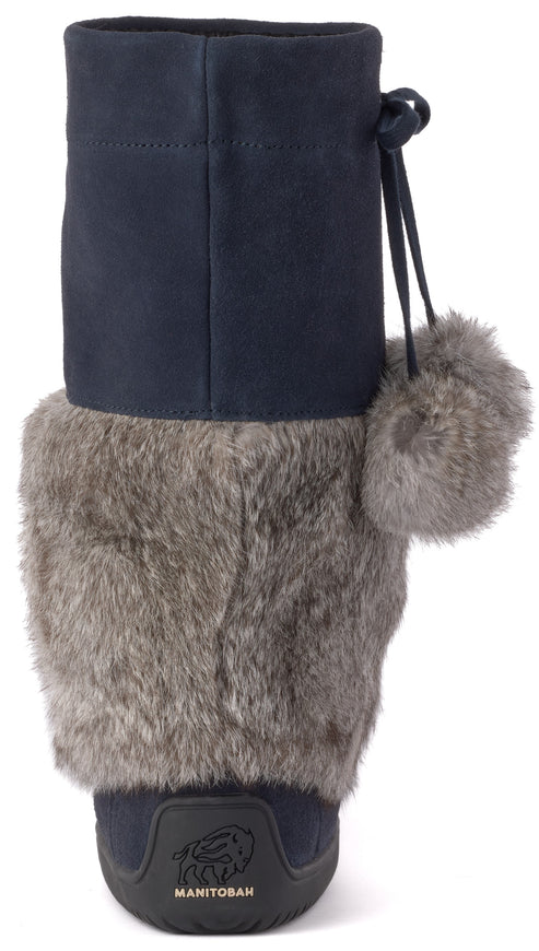 A back view of a dark blue calf-high winter boot, featuring a plush grey faux fur cuff covering the lower portion and a decorative grey pom-pom hanging from a drawstring at the top, with a Manitobah logo embossed on the dark rubber heel.