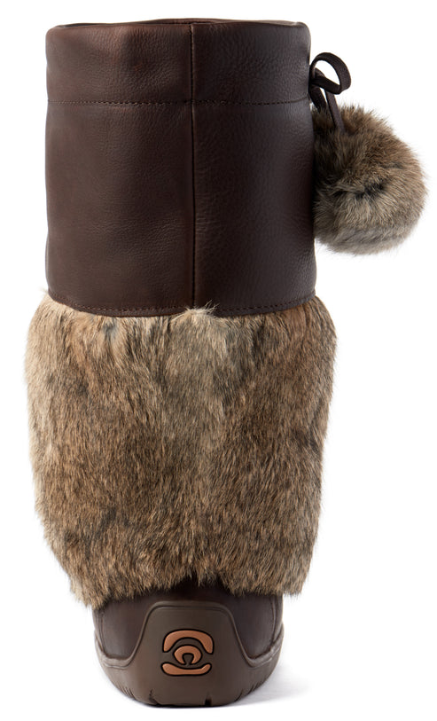 A back view of a brown leather winter boot, featuring a thick, shaggy brown faux fur trim around the mid-section, a decorative fur pom-pom hanging from the top side, and a brown plastic heel cap at the base with a small, stylized symbol embossed in the center.