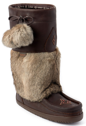 A brown winter boot with a smooth leather lower foot, a faux fur mid-shaft section, and a darker brown smooth leather upper cuff. The upper cuff features a tied leather lace with two large faux fur pom-poms dangling from the side. A small, multi-colored geometric beadwork design is stitched on the top of the toe and on the upper leather cuff.