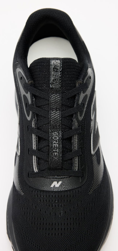 A top-down close-up view of a black athletic shoe. The shoe features a black mesh upper, a lacing system with a reflective accent strip down the center that reads GORE-TEX, and a small New Balance N logo on the base of the laces.