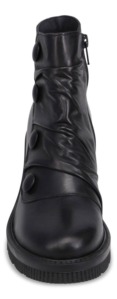 A black ankle boot with a rounded toe, a thick ridged lug sole, and a decorative strap on the side adorned with four black circular buttons, featuring a zippered closure on the inner side.