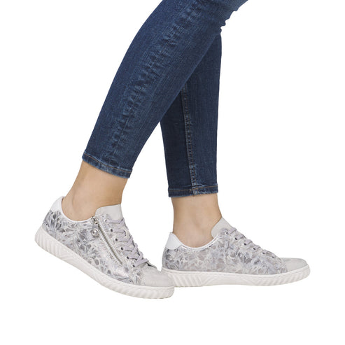 A person wearing silver floral patterned sneakers with a side zipper and blue jeans.