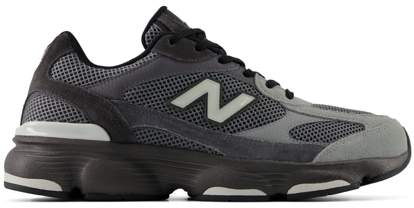 A New Balance 990v6 running shoe in a grey and black colorway featuring a mesh and suede upper, black laces, and the signature white letter N on the side, set against a chunky, dark-colored foam midsole with white rubber outsole accents.