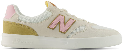 A low-top sneaker featuring an off-white upper with a pink logo on the side, a mustard yellow suede heel panel, a light pink heel tab, and a white sole.