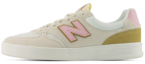 Side view of a low-top athletic shoe in a light cream color, featuring a soft pink 'N' logo outlined in gold, a yellow-ochre suede heel patch with a small pink trim at the top, and a flat white sole.