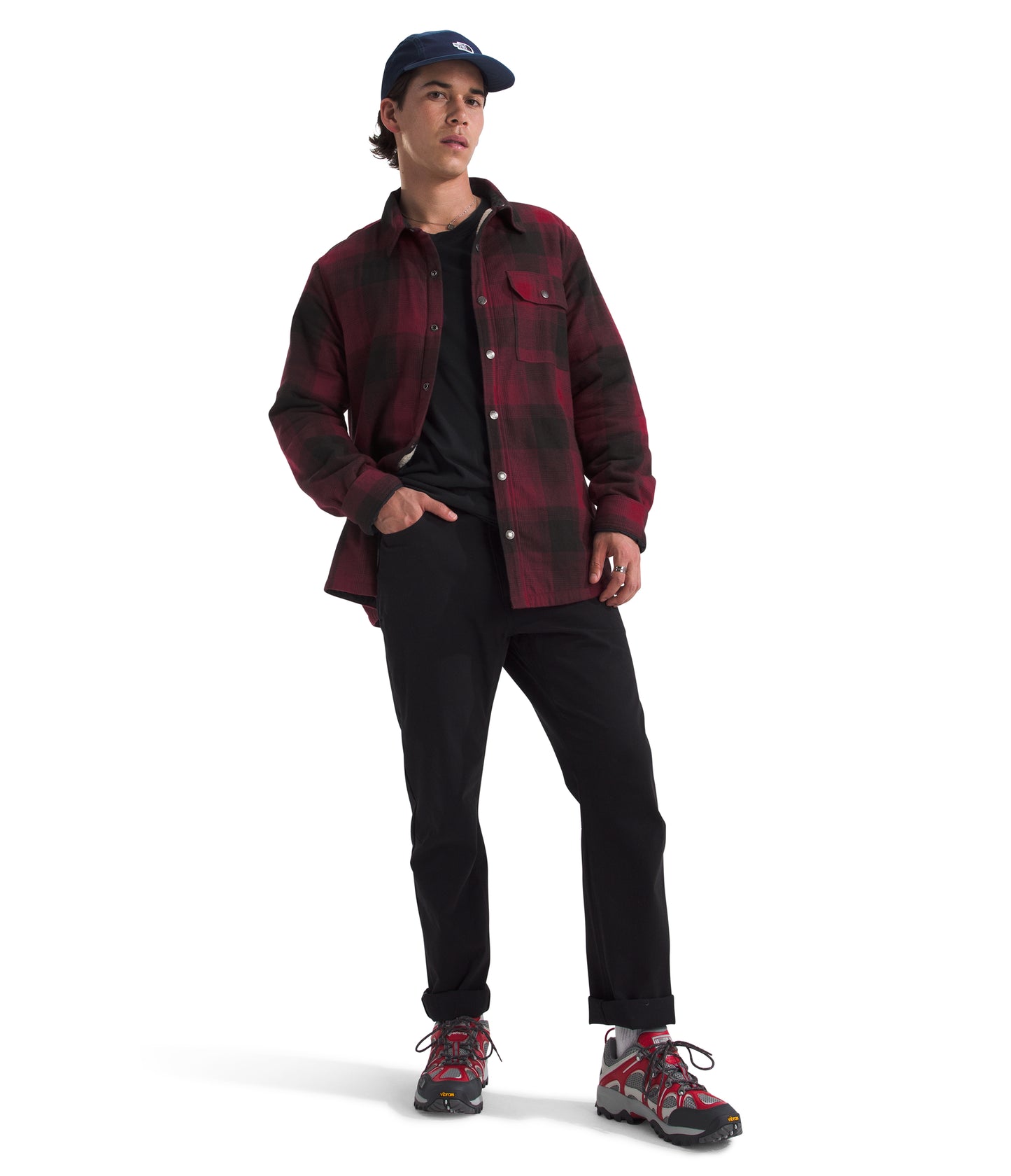 A man in a blue baseball cap, black t-shirt, black jeans, and red and black plaid jacket with red and black hiking boots.