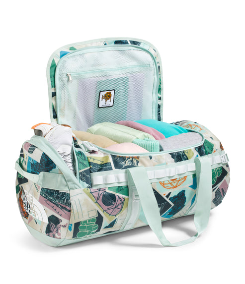 Open duffel bag in patchwork pattern with light green handles, showing packed clothing and a zippered mesh compartment inside the lid.