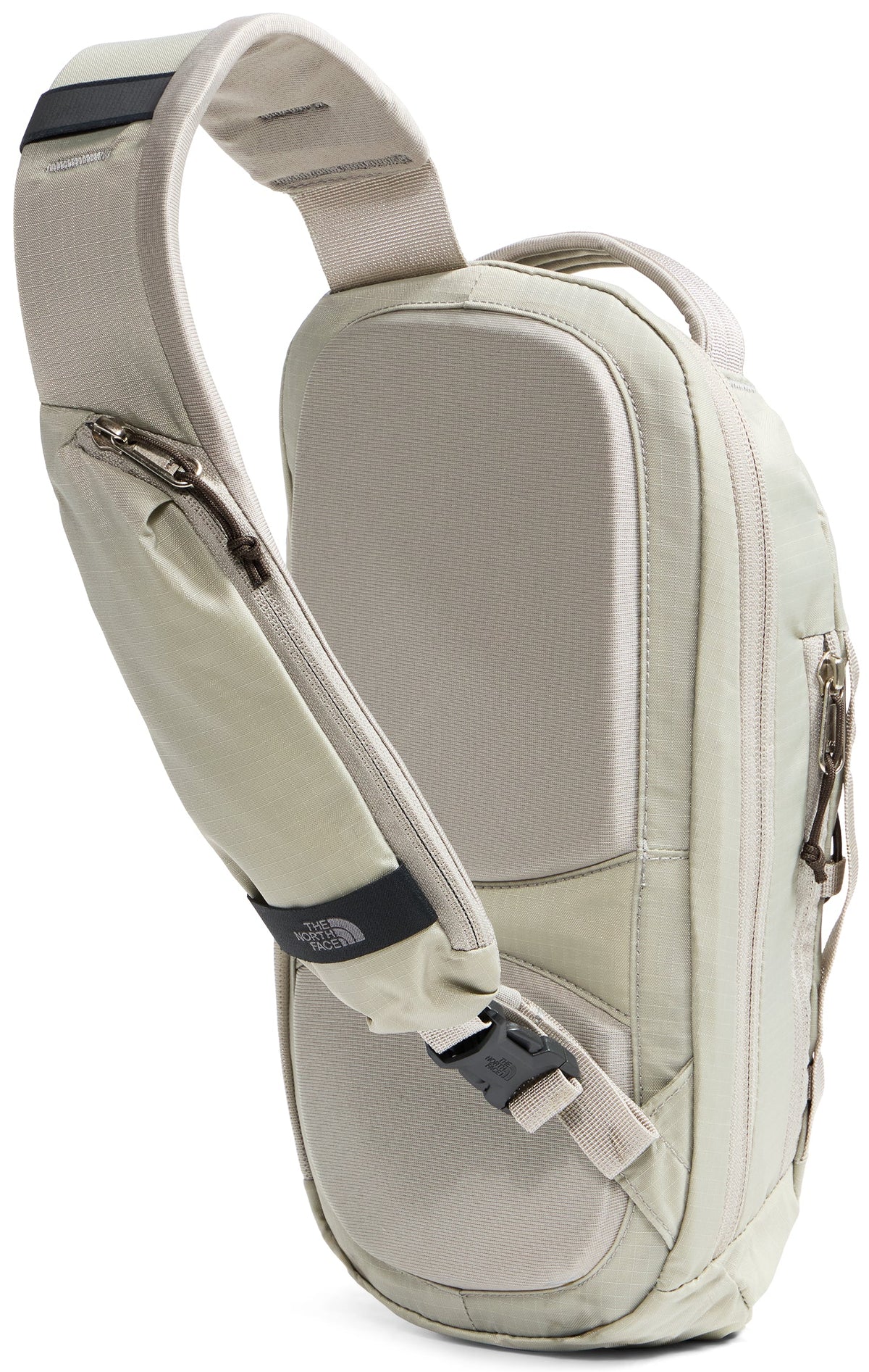 A light beige, single-strap sling backpack shown from the back, featuring a padded back panel with mesh texture, a main zippered compartment, and a contoured shoulder strap with a zippered pocket, a black plastic buckle, and a small black rectangular brand label.