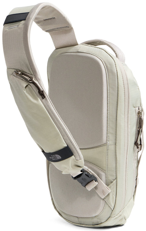A light beige, single-strap sling backpack shown from the back, featuring a padded back panel with mesh texture, a main zippered compartment, and a contoured shoulder strap with a zippered pocket, a black plastic buckle, and a small black rectangular brand label.