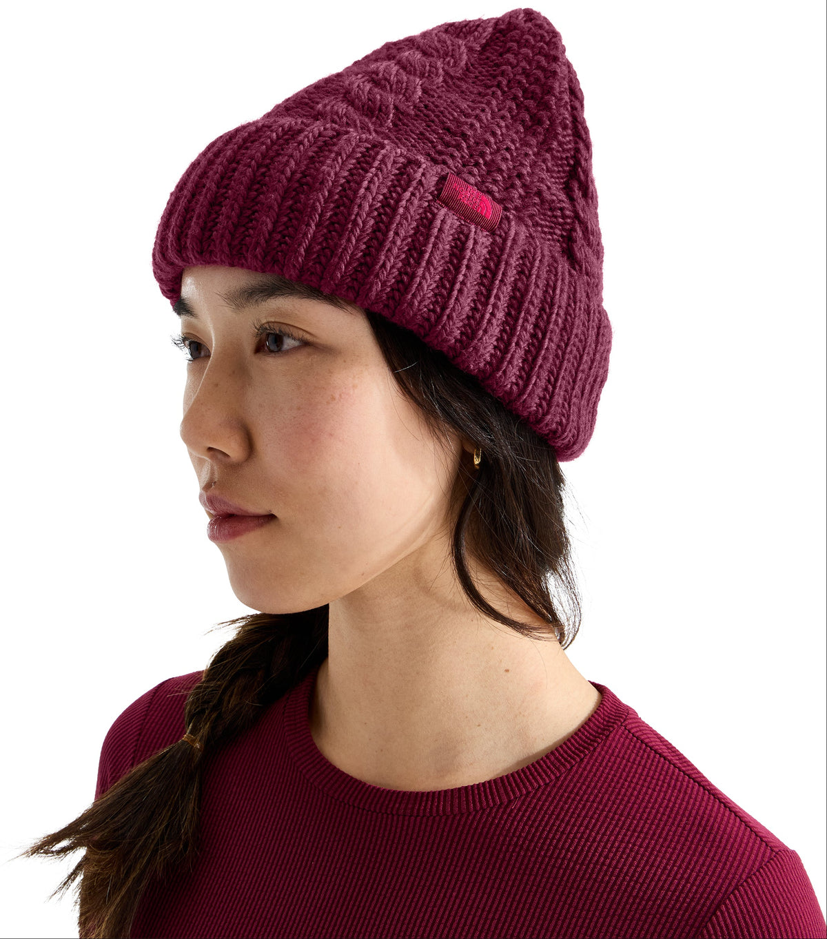 A woman wearing a dark burgundy cable-knit winter beanie with a ribbed cuff and a small square tag on the side, paired with a matching long-sleeved burgundy ribbed shirt.