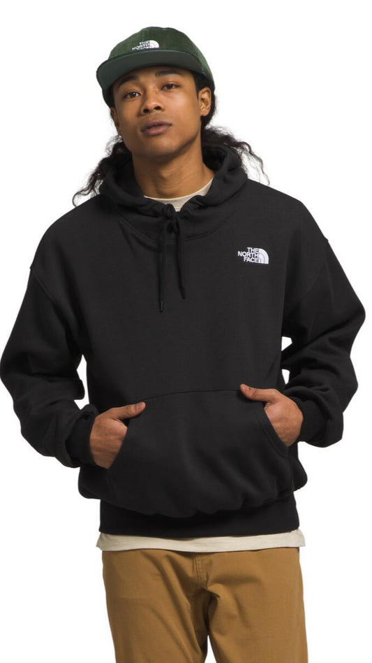 Man wearing a dark green cap, black hoodie, and tan pants stands with hands in hoodie pocket. "North Face" logo is visible on the hoodie.