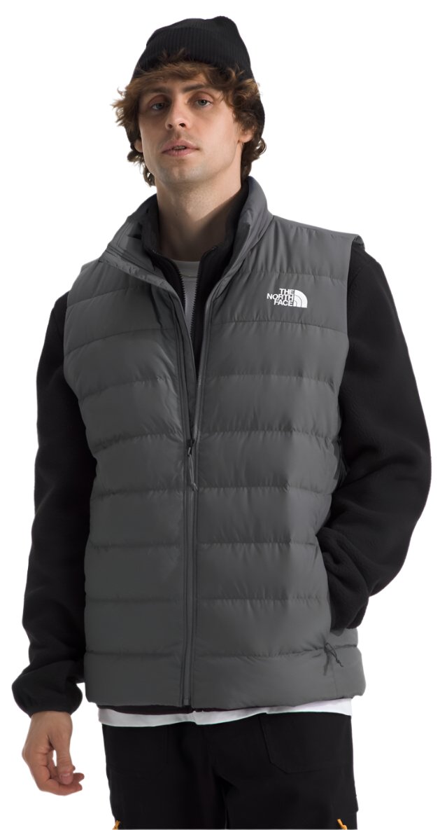 A person wears a gray puffy vest with a white logo on the left side of the chest and a black hat.