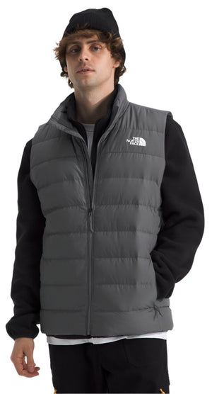 A person wears a gray puffy vest with a white logo on the left side of the chest and a black hat.