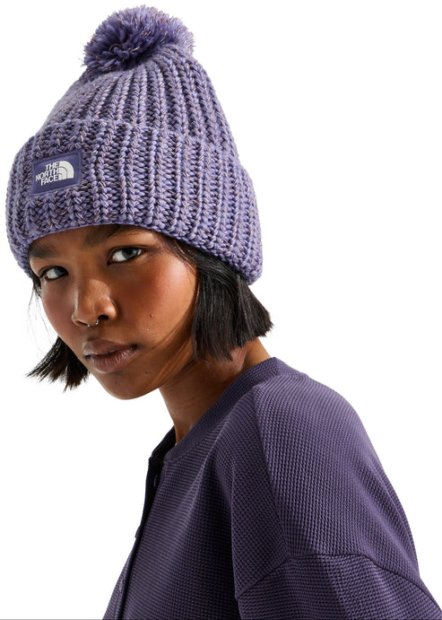 A person wearing a purple, chunky knit, ribbed beanie with a matching pom-pom on top and a square logo patch on the folded cuff, paired with a matching purple waffle-knit long-sleeve top.