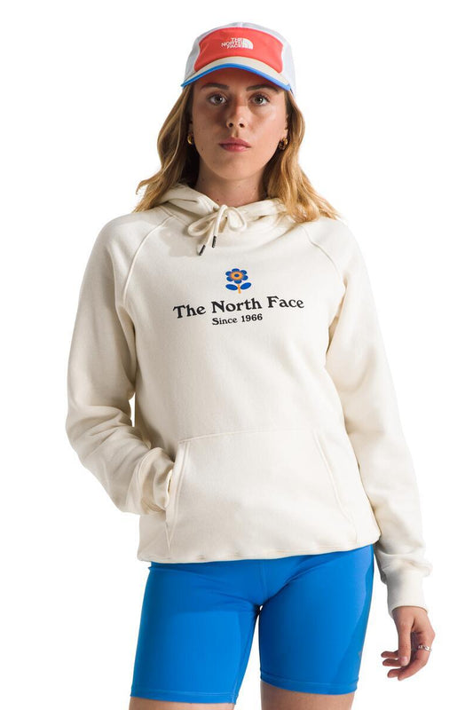 A woman wearing a cream-colored hoodie with "The North Face Since 1966" printed on the chest with a small flower design above the text, paired with blue shorts and a white and orange hat.