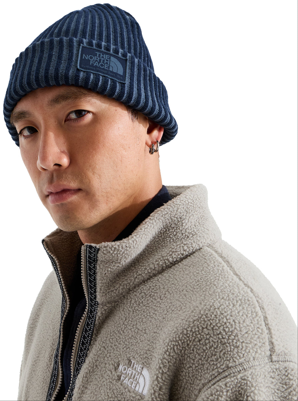 A man wearing a navy blue ribbed knit beanie with a rectangular patch featuring text, and a light beige, textured fleece zip-up jacket with an embroidered logo on the chest.