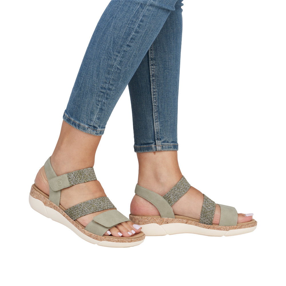 A person is modeling pale green, open toe sandals with elastic straps and a white sole while wearing jeans.