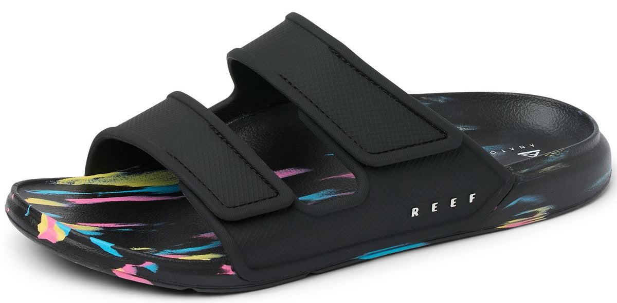 A black sandal with two velcro straps over the foot and a colorful paint-splattered sole; the word "REEF" is printed in white letters on the side.