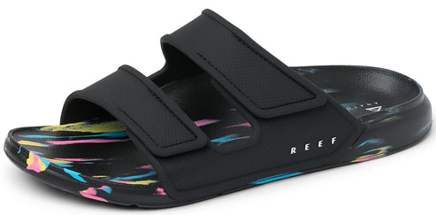 A black sandal with two velcro straps over the foot and a colorful paint-splattered sole; the word "REEF" is printed in white letters on the side.