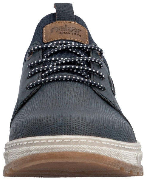 A low-angle front view of a dark navy blue casual men's sneaker featuring a textured synthetic upper, a brown leather-like tongue patch with the text 'rieker since 1874', speckled black-and-white laces, and a thick, ridged white midsole with a gum-colored rubber outsole.