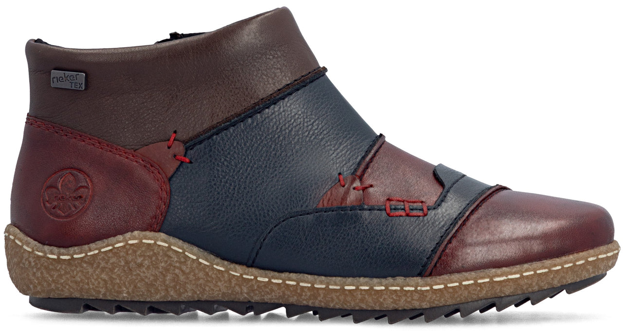 A leather ankle boot with a multi-colored patchwork design, featuring segments of dark red, navy blue, and brown, accented with red stitching, a textured tan rubber sole, and a Rieker logo embossed on the heel.