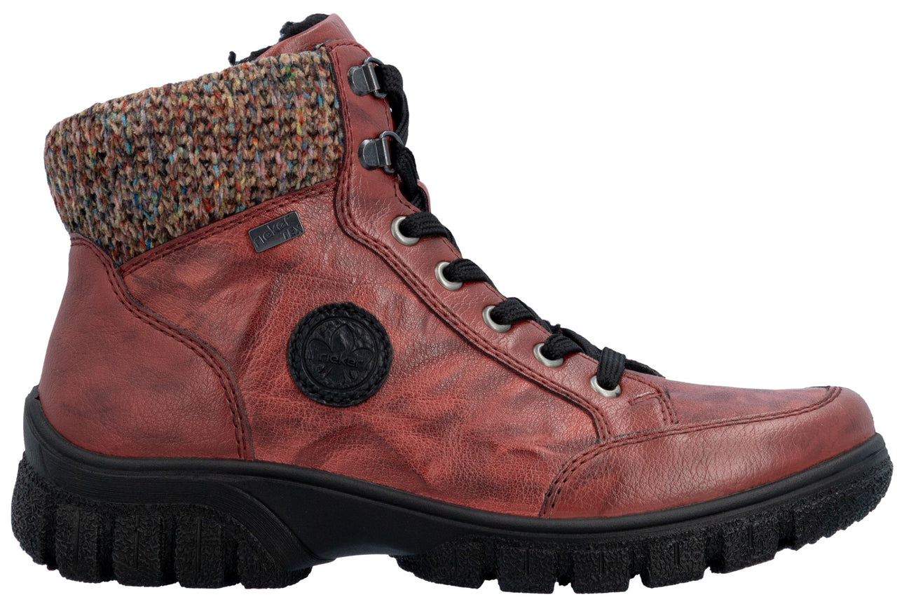 A red ankle boot with a multi-colored knit cuff at the collar, a black lace-up closure with metallic eyelets, and a thick, ridged black rubber lug sole, featuring a circular black logo patch on the side.