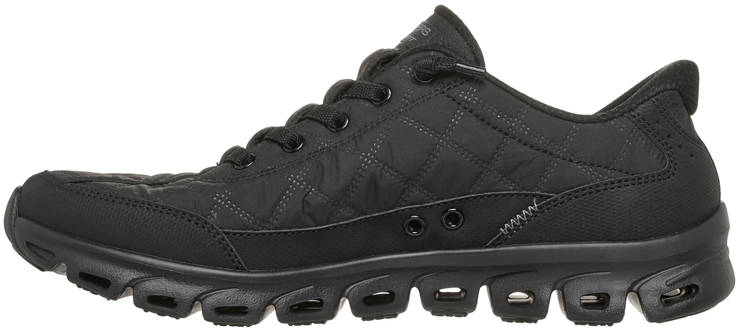A black quilted low-top athletic shoe with black laces and a thick, segmented black rubber sole featuring open cut-outs for flexibility.