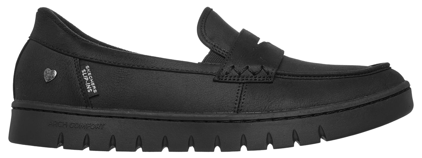 A black slip-on loafer with a thick, serrated outsole, a heart-shaped emblem on the heel, and a small fabric tag reading "SKECHERS SLIP-INS" on the side.