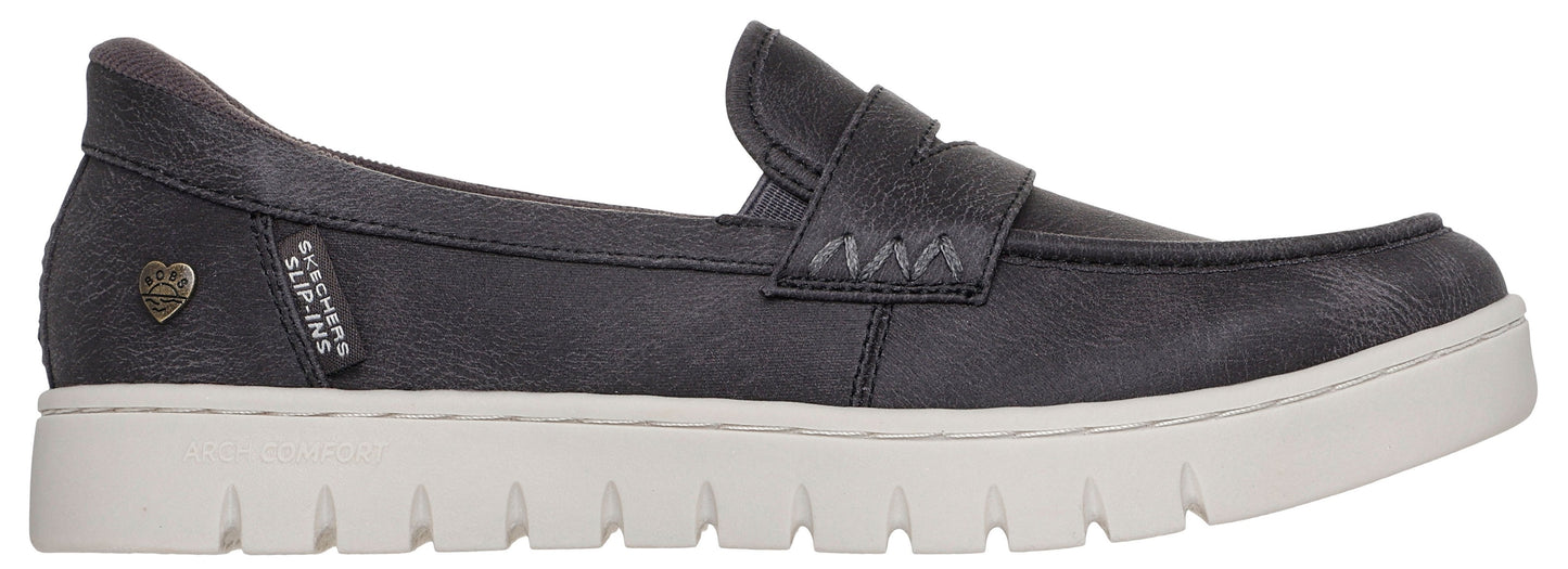 A black slip-on loafer with a thick, textured white sole, featuring a small metallic heart emblem and a small fabric tag reading 'SKECHERS SLIP-INS' on the side.