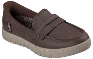 A brown slip-on loafer shoe with a thick, textured beige rubber sole, featuring a saddle strap across the upper and a small tag on the side that reads SKECHERS SLIP-INS.