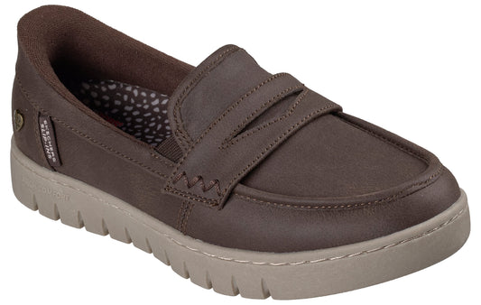 A brown slip-on loafer shoe with a thick, textured beige rubber sole, featuring a saddle strap across the upper and a small tag on the side that reads SKECHERS SLIP-INS.