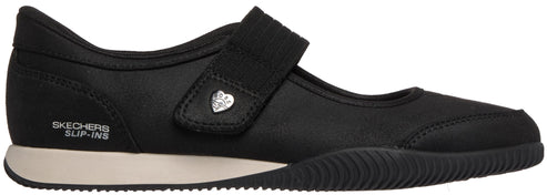 A black Mary Jane-style slip-on shoe with a rounded toe, a single hook-and-loop strap featuring a small heart-shaped embellishment, a white midsole, and a textured black outsole, with the text SKECHERS SLIP-INS visible on the side heel.