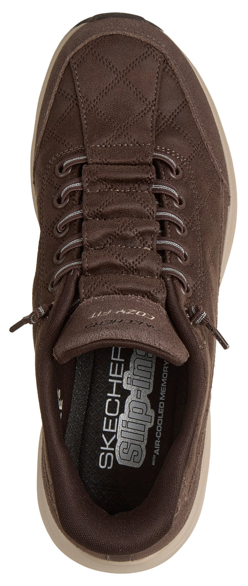A top-down view of a dark brown slip-on shoe with a quilted upper texture, featuring decorative laces, a cushioned interior with text branding, and a light-colored sole.