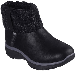 A black ankle boot with a smooth, matte-textured lower section and a thick, chunky knit sweater-style cuff at the top. The boot has a thick, dark gray midsole and a black treaded outsole, with a small vertical tag reading "SKECHERS" on the side.