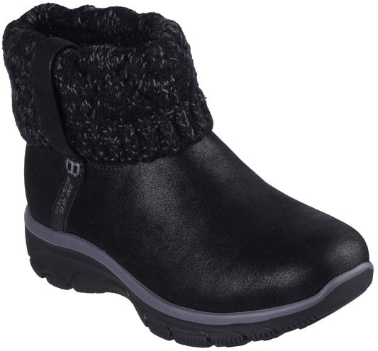 A black ankle boot with a smooth, matte-textured lower section and a thick, chunky knit sweater-style cuff at the top. The boot has a thick, dark gray midsole and a black treaded outsole, with a small vertical tag reading "SKECHERS" on the side.