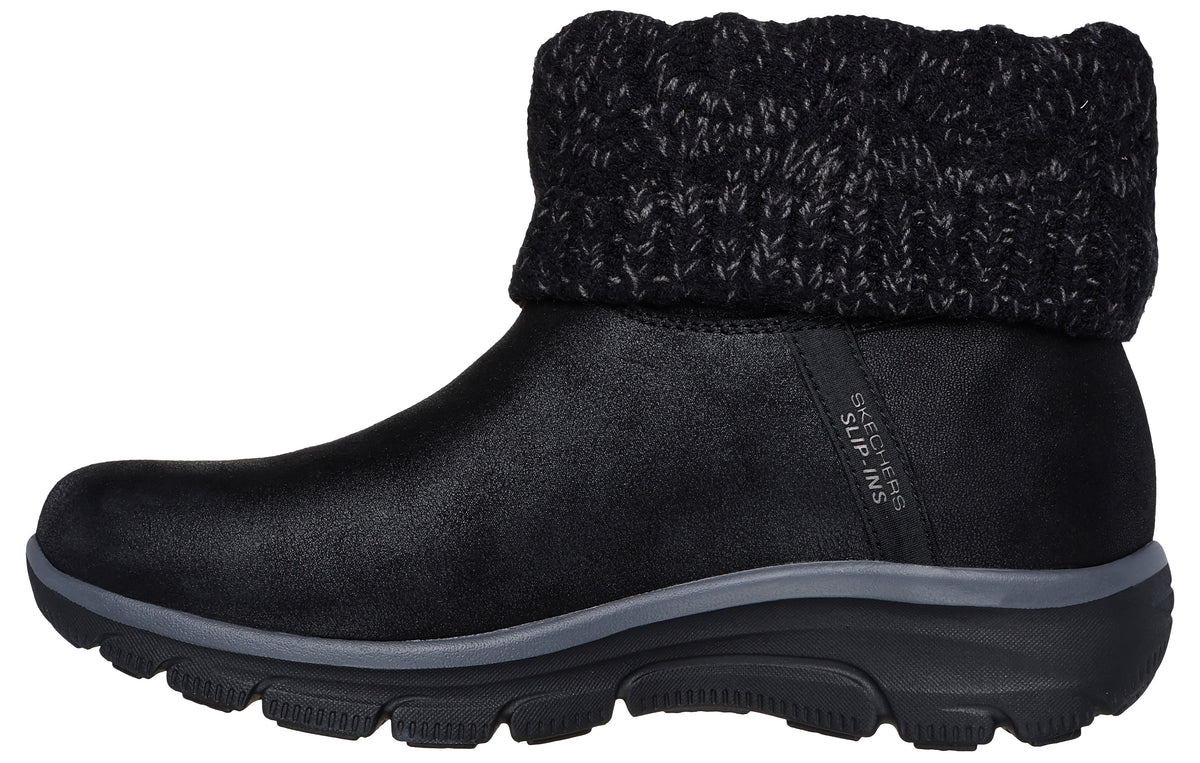 A black slip-on ankle boot with a black knit sweater-style cuff, a smooth matte black upper with "SKECHERS SLIP-INS" branding on the side, and a grey and black cushioned sole.