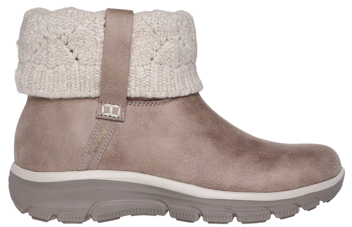 A tan faux-suede ankle boot with a beige, knit, sweater-like cuff around the ankle, secured by a small matching strap, set on a thick, rugged, light brown rubber sole.