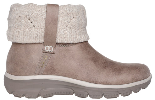 A tan faux-suede ankle boot with a beige, knit, sweater-like cuff around the ankle, secured by a small matching strap, set on a thick, rugged, light brown rubber sole.