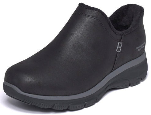 A black, slip-on ankle boot with a rounded toe, a grey midsole, a black lugged outsole, and a soft, dark-colored faux fur lining along the collar, with the text 'SKECHERS SLIP-INS' visible near the heel.