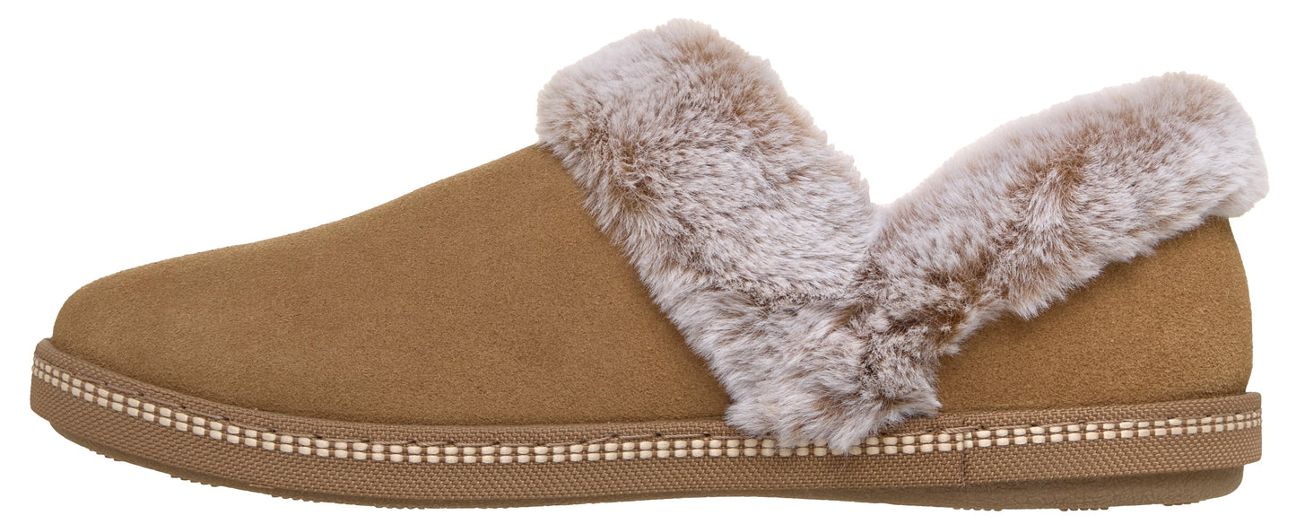 A tan suede slip-on slipper with a brown rubber sole that has decorative stitching and a light brown faux fur trim around the opening.