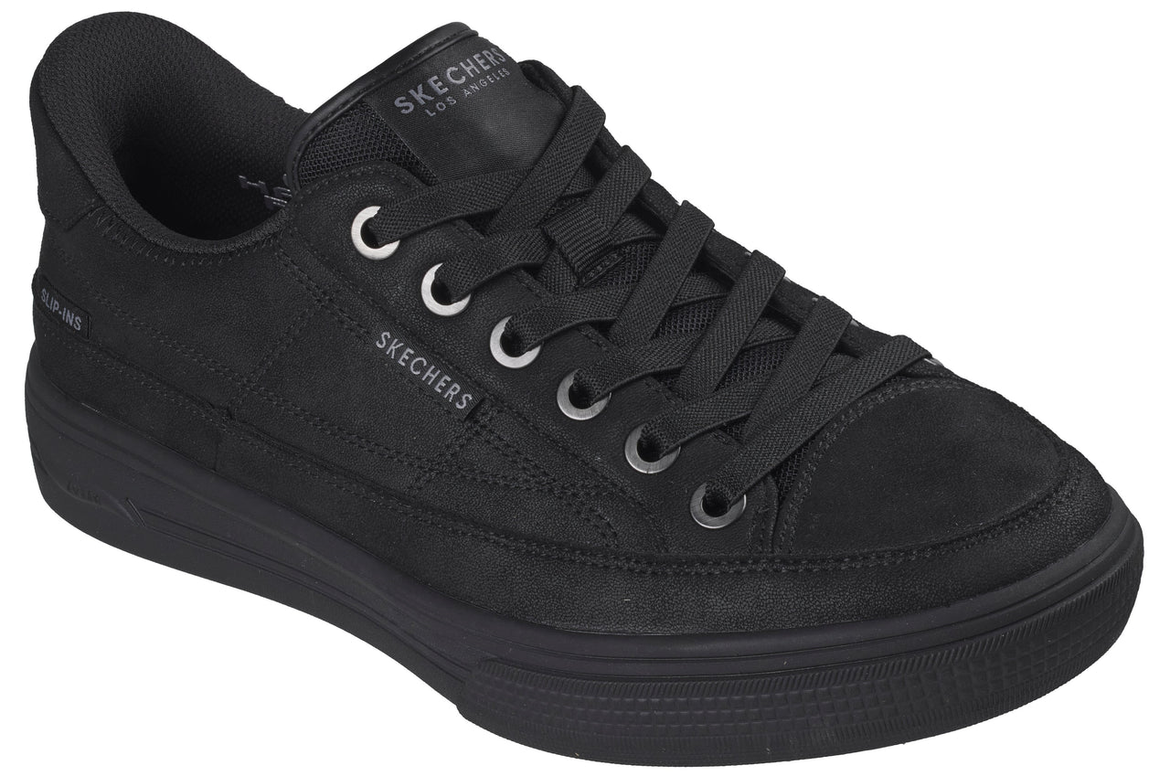 A low-top, all-black sneaker with a canvas upper, a thick rubber midsole, and matching black laces threaded through silver-toned eyelets, featuring the brand name visible on the tongue and side.