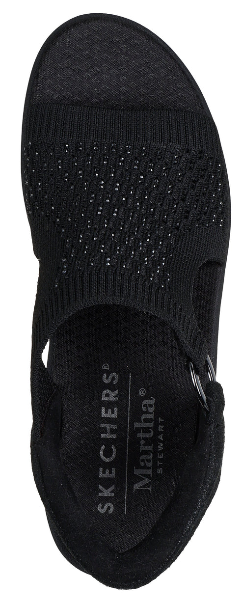 A black sandal with an elastic strap and a flat sole. The insole has white text on it that reads "SKECHERS" and "Martha Stewart".