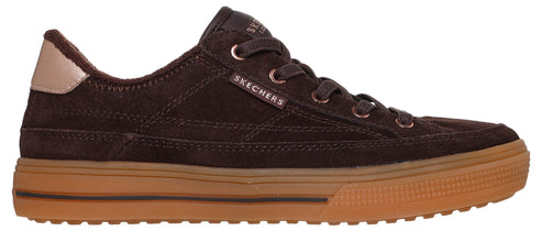 A brown suede low-top sneaker featuring a thick, textured gum rubber sole, brown laces with rose gold eyelets, and a small rose gold Skechers logo tag on the side, with a light tan heel accent.