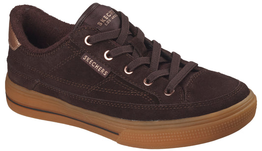 A brown suede low-top sneaker with a textured gum rubber midsole and a metallic copper-toned heel tab, featuring the brand name visible on the tongue and side, and flat brown laces.