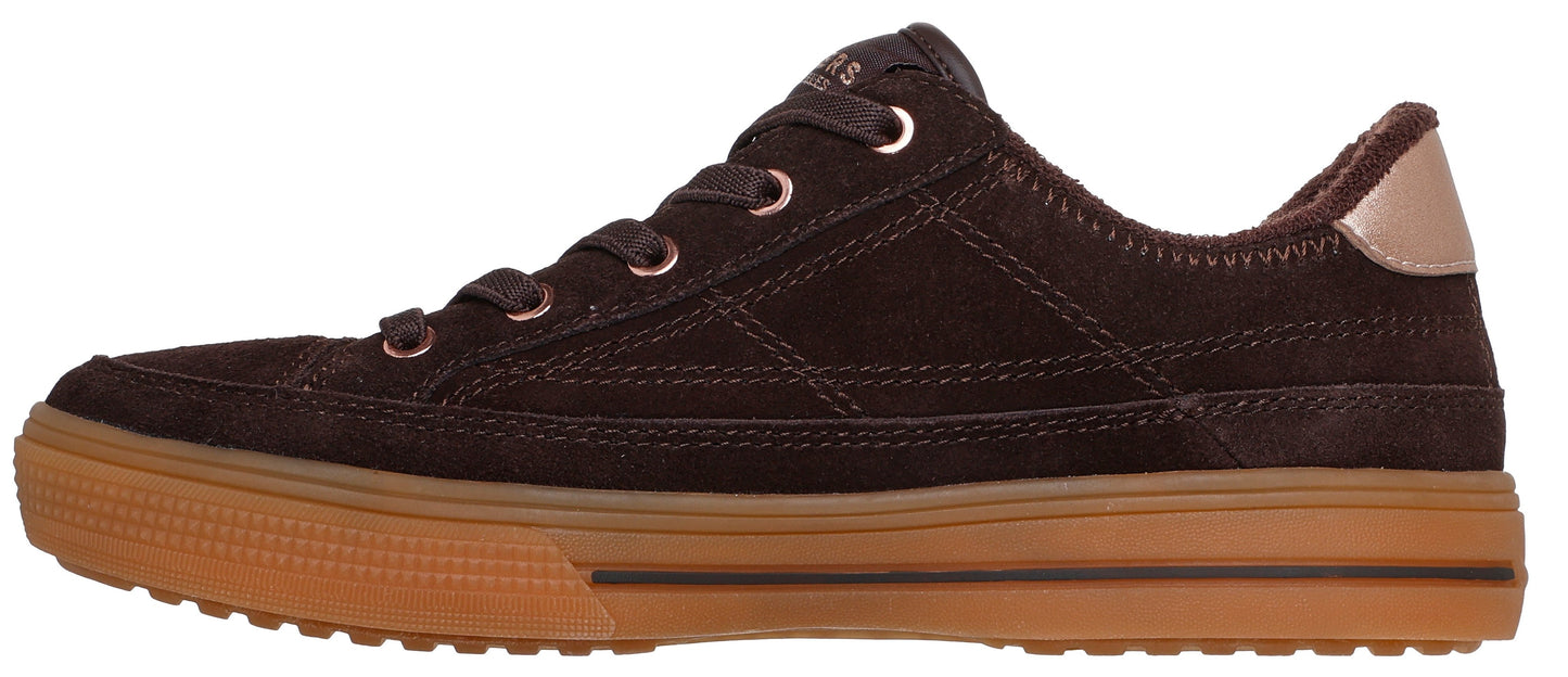 A brown, suede, lace-up sneaker with a flat gum rubber sole, a metallic rose-gold heel tab, and metallic rose-gold lace eyelets.