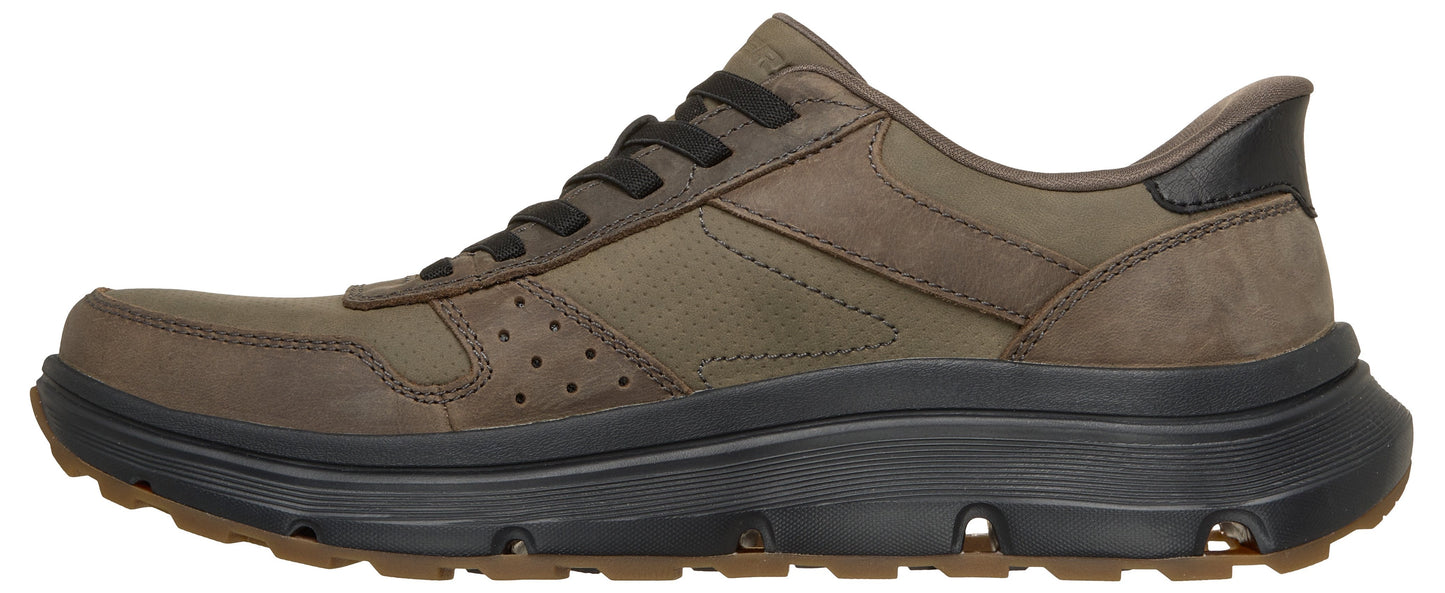 A low-top sneaker featuring an olive green nubuck leather upper with dark brown laces and a black heel accent, set atop a chunky, textured black foam midsole and a brown lugged rubber outsole.