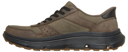 A low-top sneaker featuring an olive green nubuck leather upper with dark brown laces and a black heel accent, set atop a chunky, textured black foam midsole and a brown lugged rubber outsole.