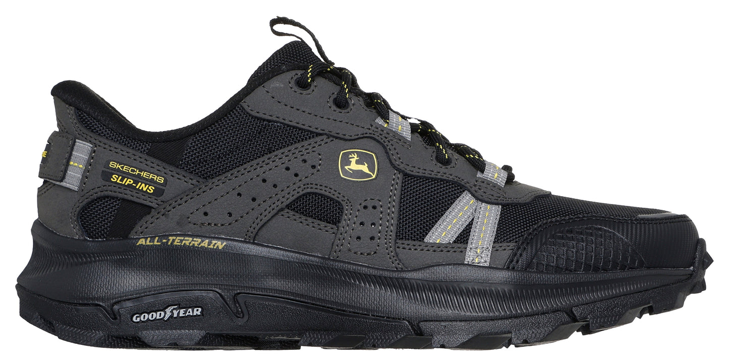 A side view of a dark gray and black athletic shoe with the Goodyear logo on the black sole and a John Deere logo on the side.