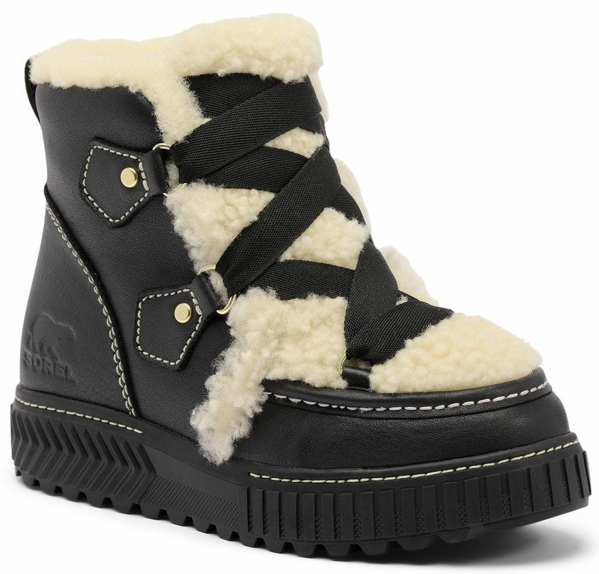 A black ankle boot with a white shearling trim, featuring crisscross black fabric lacing, gold hardware accents, a thick lugged sole, and white contrast stitching, showing the SOREL brand logo embossed on the side.